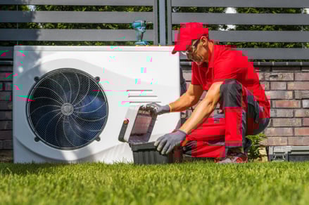 An A&E HVAC technician kneels next to an outdoor Carrier Heat Pump unit to do maintenance on.