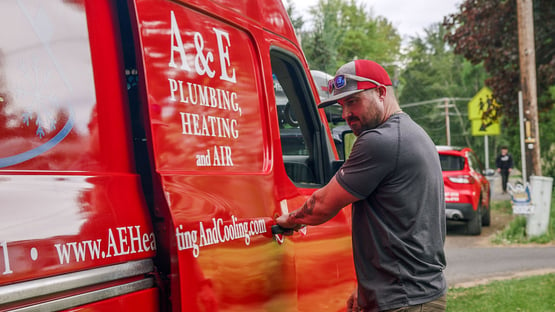 An A&E Plumbing, Heating and Air technician opening the sliding side door of a red A&E company van, presumably to grab more tools to finish an HVAC service call.