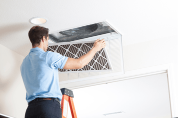 An A&E HVAC tech stands on a ladder to change an indoor HVAC air filter.
