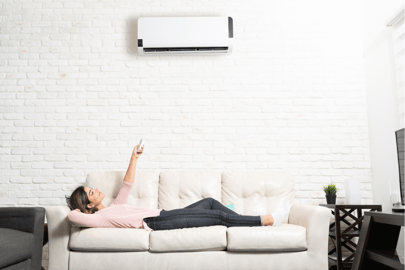 A woman lounges on a white couch, a remote pointed up at her wall-mounted mini split unit to ensure the perfect temperature in her home.