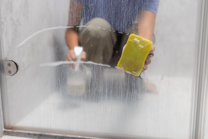 A person kneels in a shower to wipe away the hard water stains on the inside of a glass shower door.