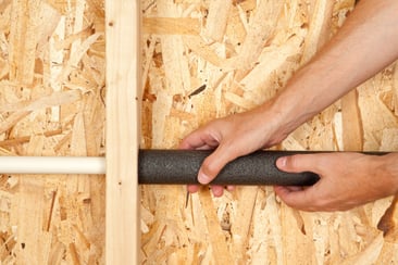 A set of hands places foam insulation around a home plumbing PVC pipe.