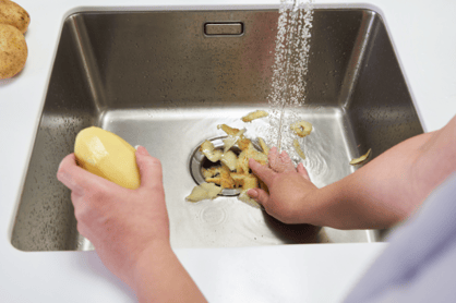 A hand pushes potato peels into a residential garbage disposal in the middle of a kitchen sink.
