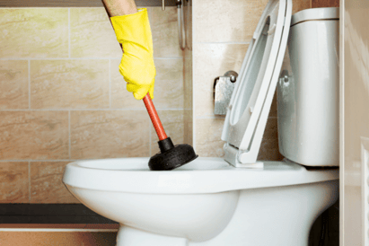 A set of yellow-gloved hands hold a plunger, inserting it into a toilet to work out a clog.