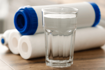 A glass of clear water sits in front of traditional water softener cartridges.