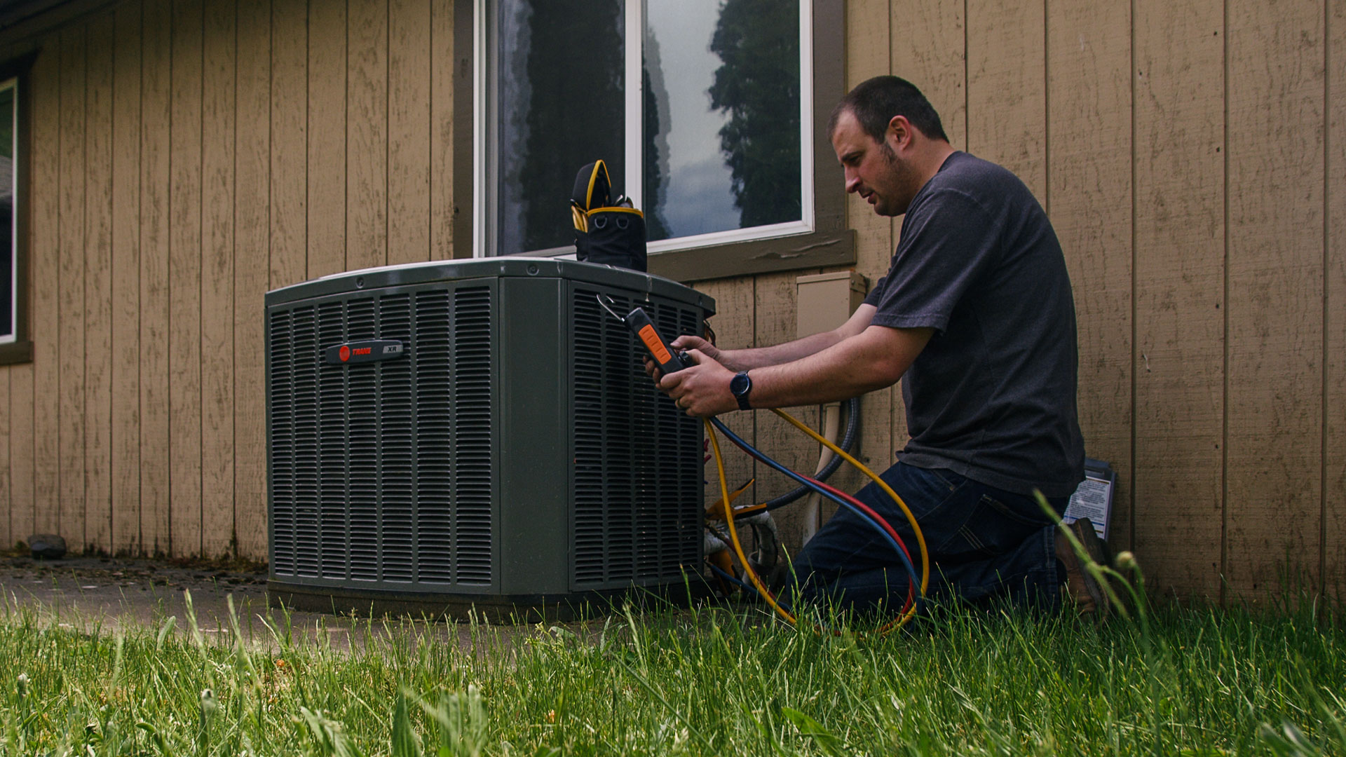 A&E HVAC tech kneeling in grass, working on outdoor unit