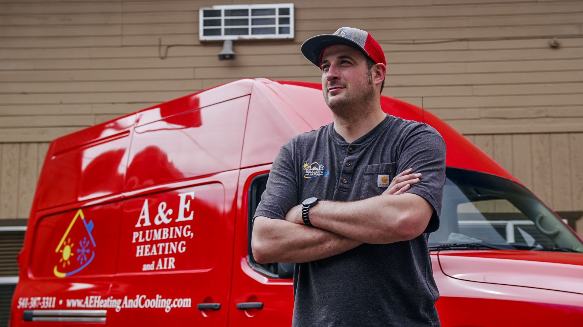 A&E tech standing in front of a bright red A&E company van, his arms folded and a small smile on his face.